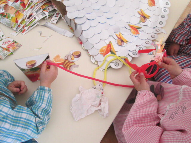 Execução de uma máscara com os frutos recortados colados nos óculos de plástico usados na casinha das bonecas e uma carteira feita com a embalagem Tetra Pak Compal e alça de trapilho.