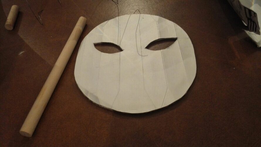 Parte frontal da máscara feita com embalagem Tetra Park da marca Compal e cilindro de madeira para suporte da máscara.